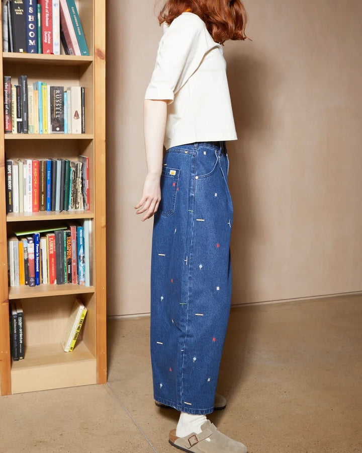 A person standing in a room with a bookshelf, wearing a white cropped short-sleeve top, a long blue denim skirt with small colorful embroidered details, white socks, and brown slip-on shoes.